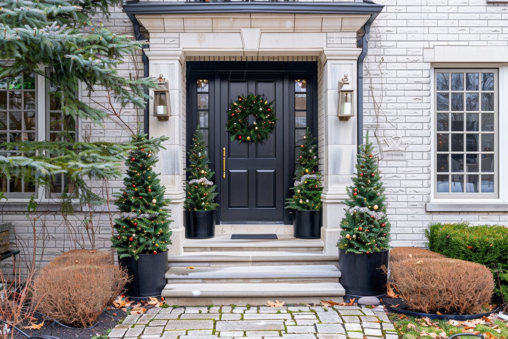 Festive porch with red door, garland, and wreath. White pillars and railing; windows with Christmas trees inside.