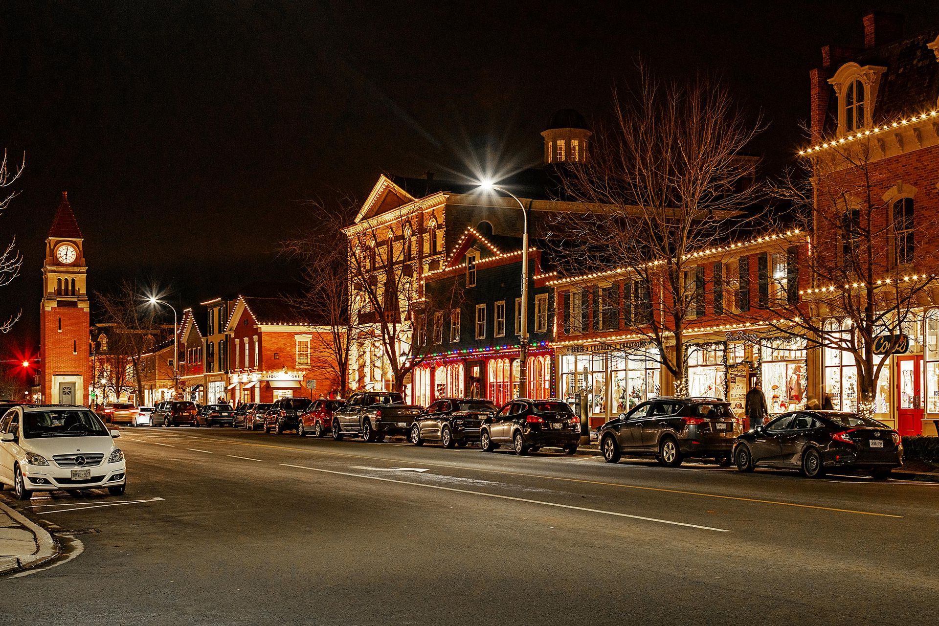 Nighttime street scene with Christmas lights, parked cars, and brick buildings.