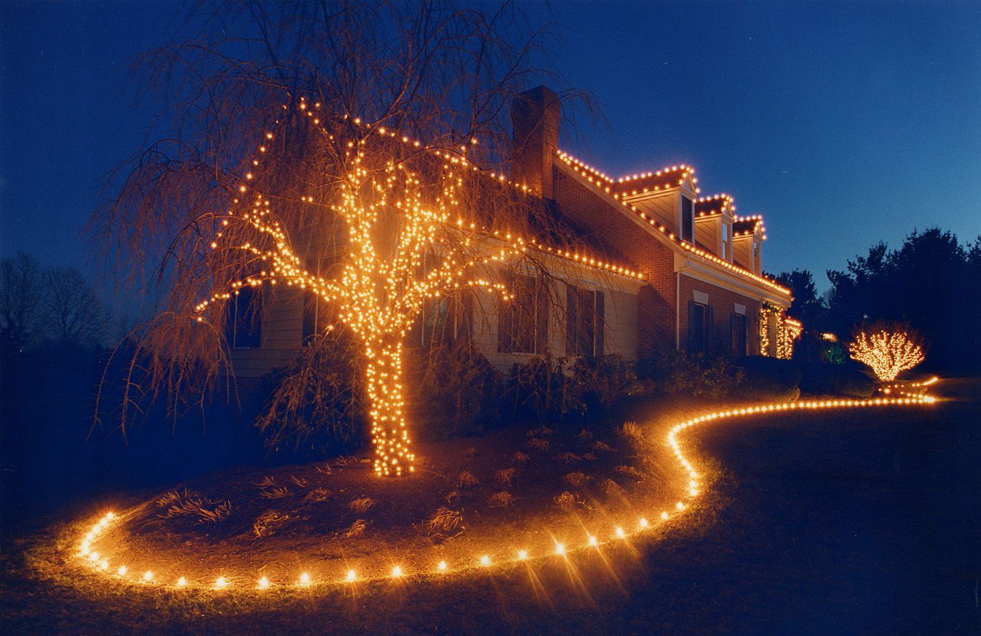 House decorated with warm white Christmas lights. Tree and walkway also lit. Nighttime scene.