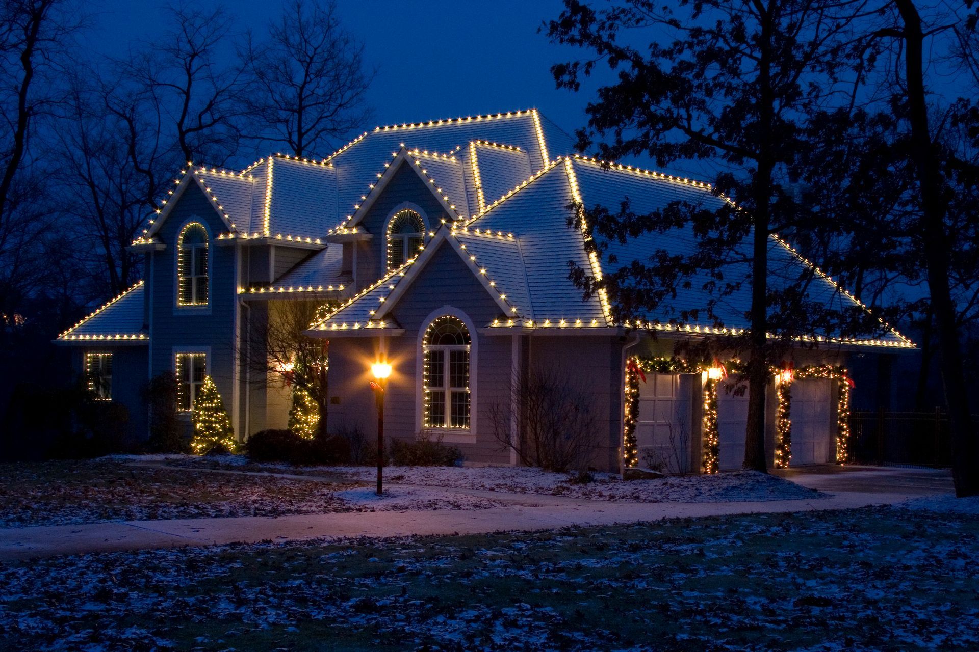 House decorated with white Christmas lights, snowy yard, dark blue sky.