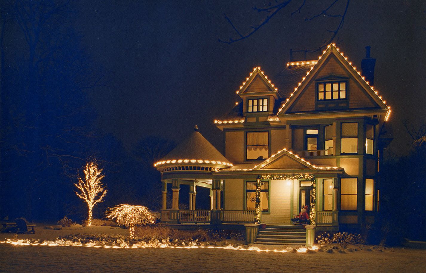 Victorian house at night, lit with Christmas lights; snow on the ground.