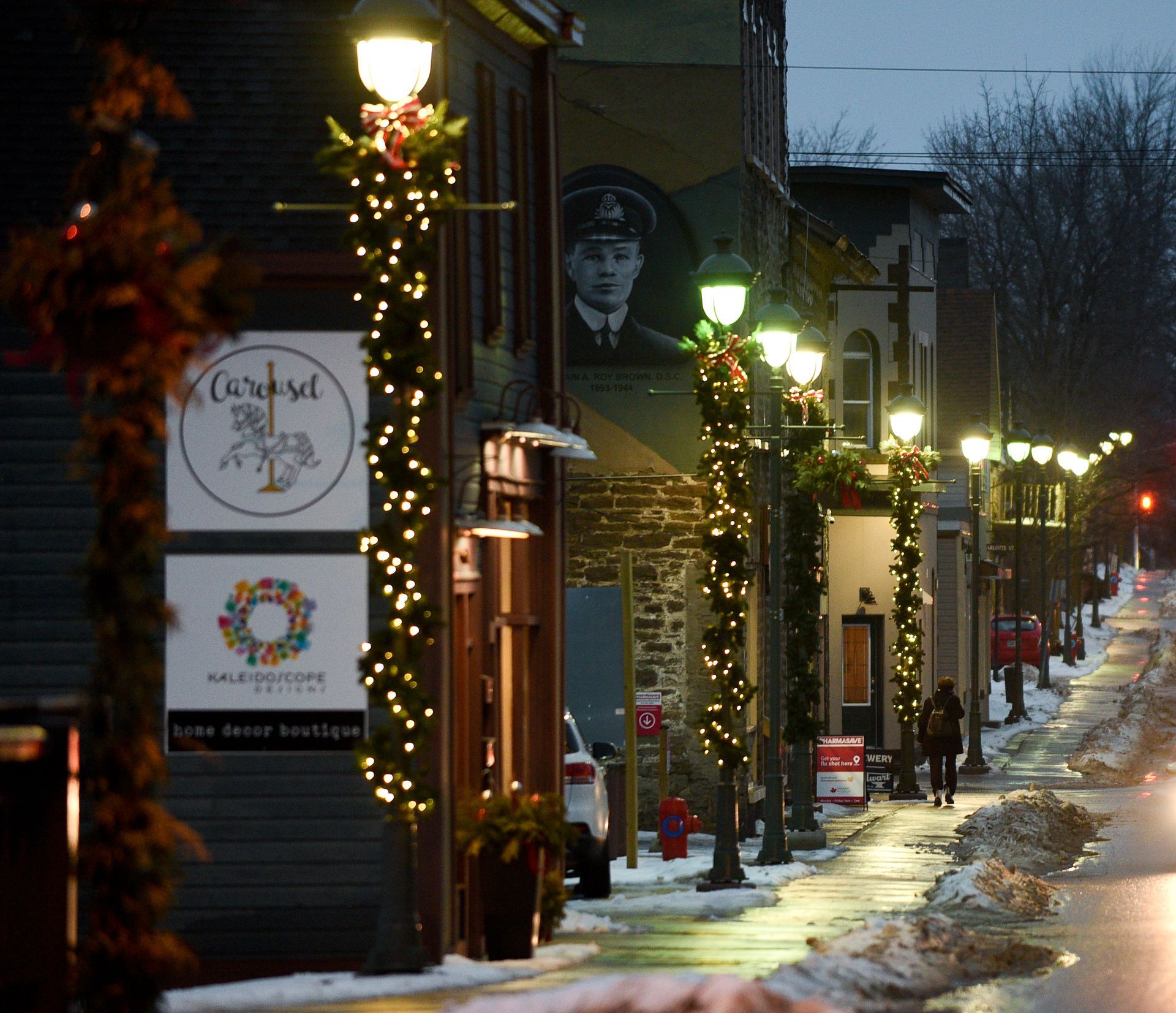Street at night with buildings and storefronts. Decorated with Christmas lights. Snowy sidewalk.