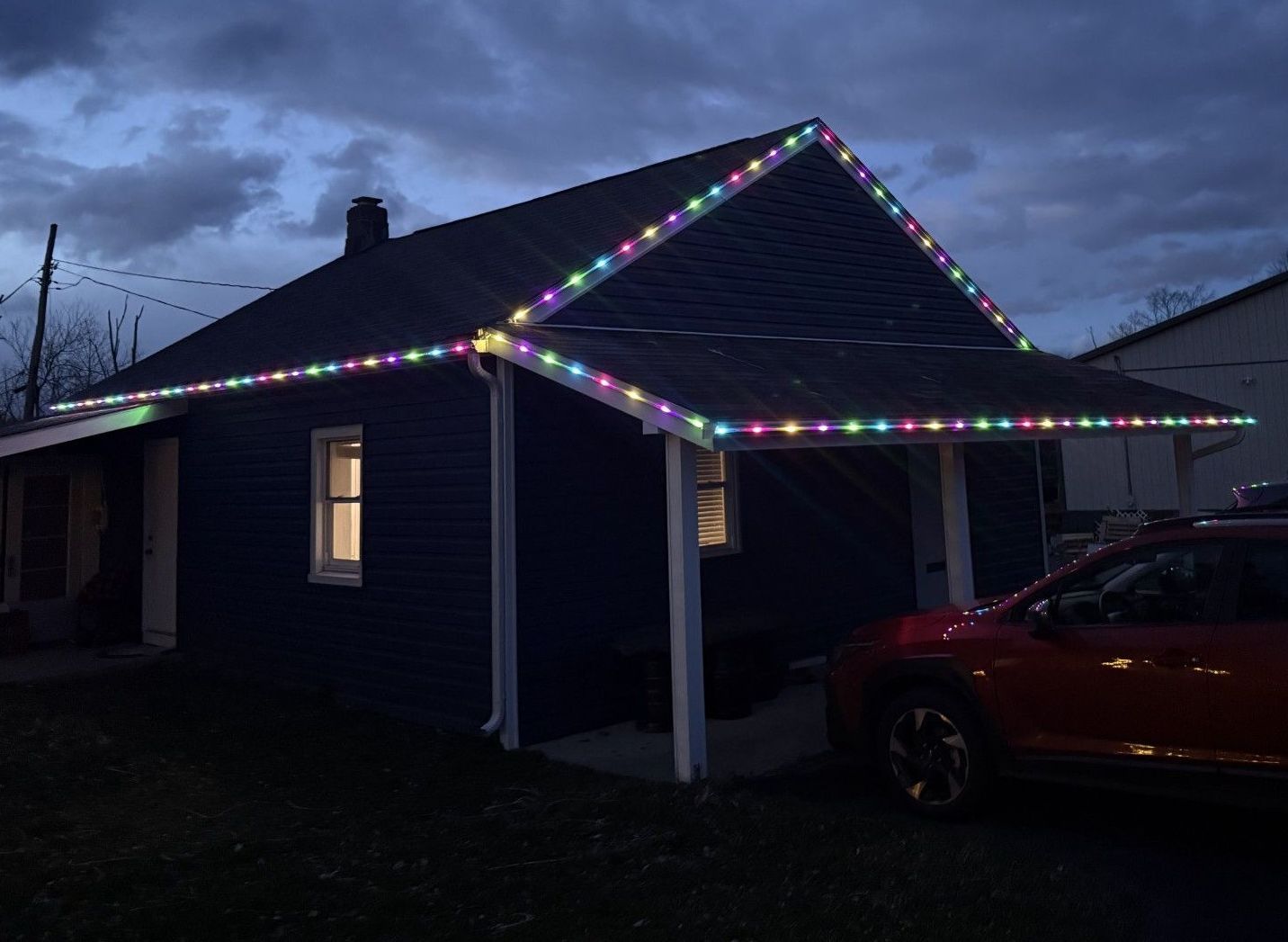 A dark blue house with colorful Christmas lights outlining the roof, and a red car parked nearby.