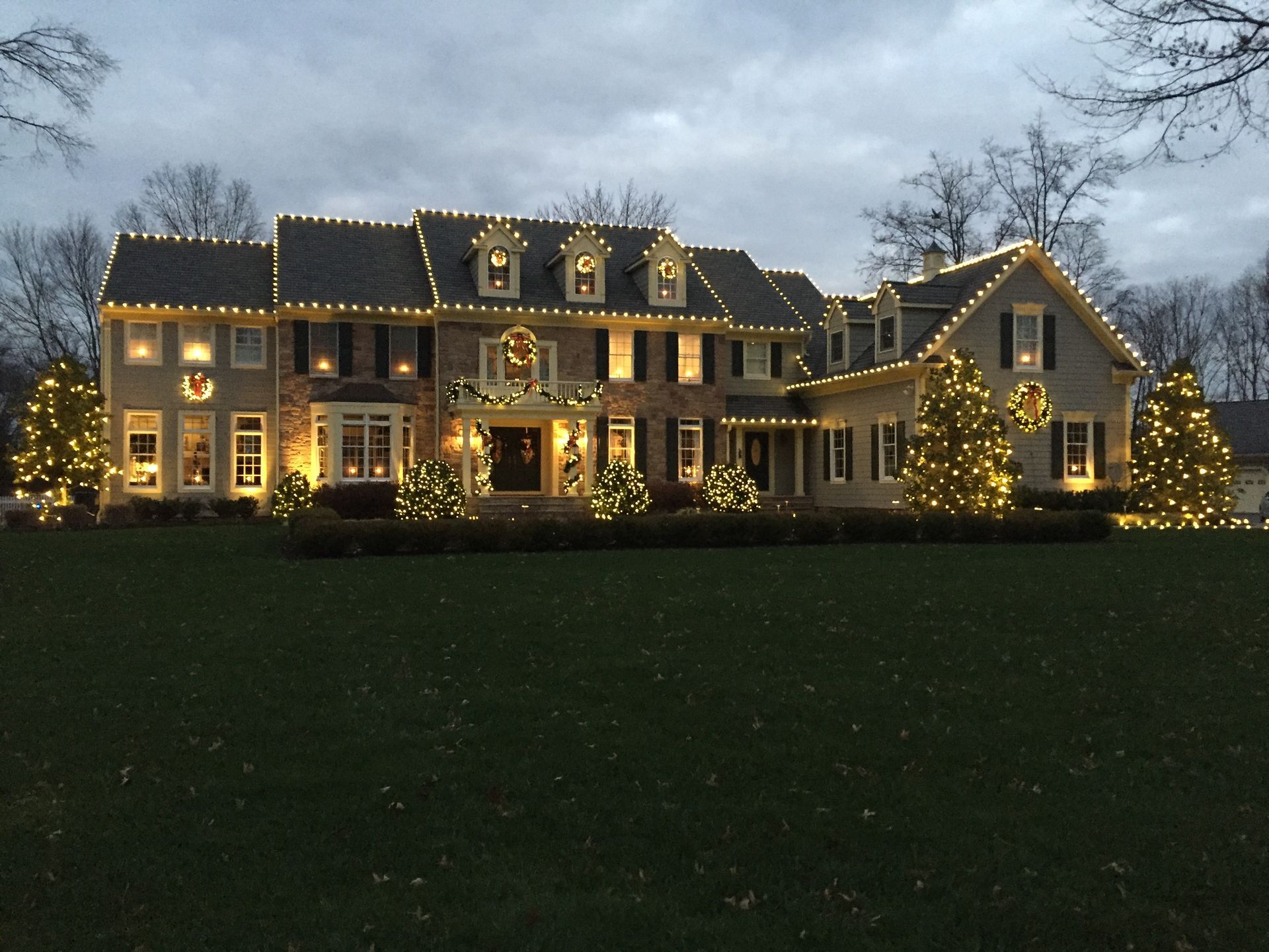 Large house decorated with Christmas lights, trees, and wreaths; evening setting.