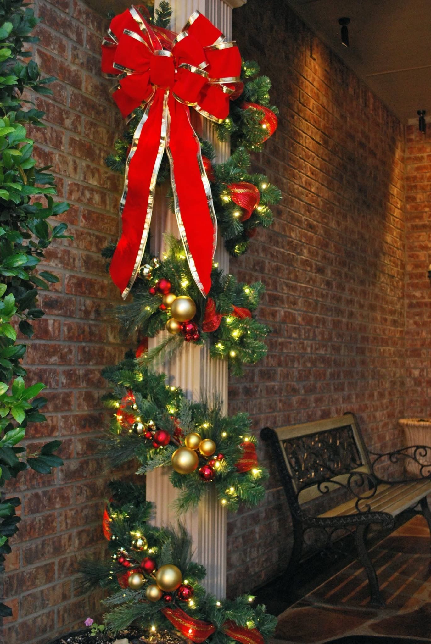 Christmas garland on a brick column, featuring a red bow and gold ornaments, with a bench in the background.