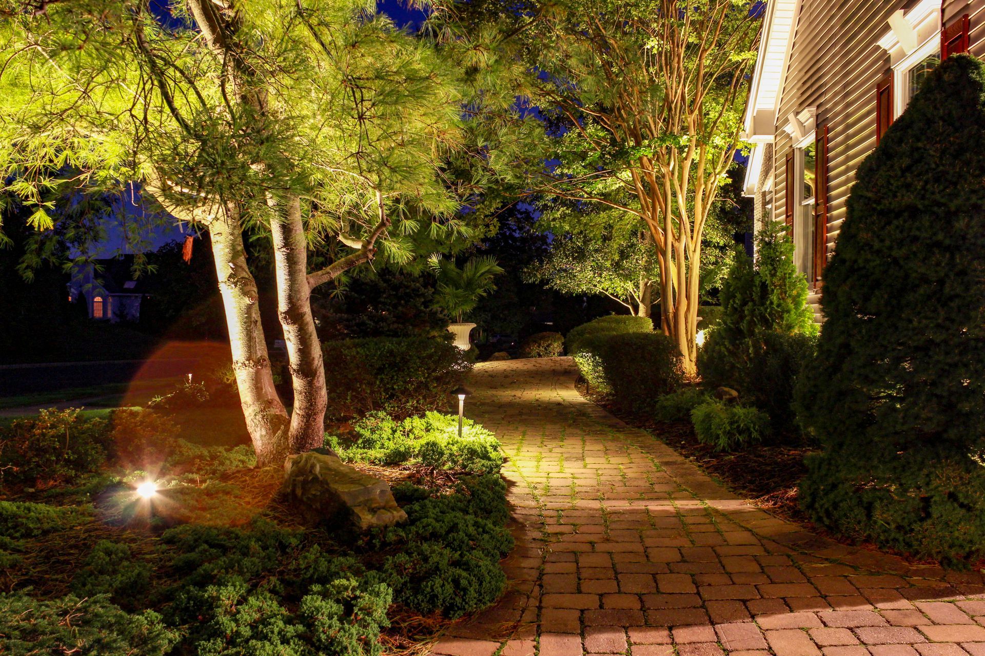 Brick pathway illuminated by spotlights, flanked by shrubs and trees, leading to a building at night.
