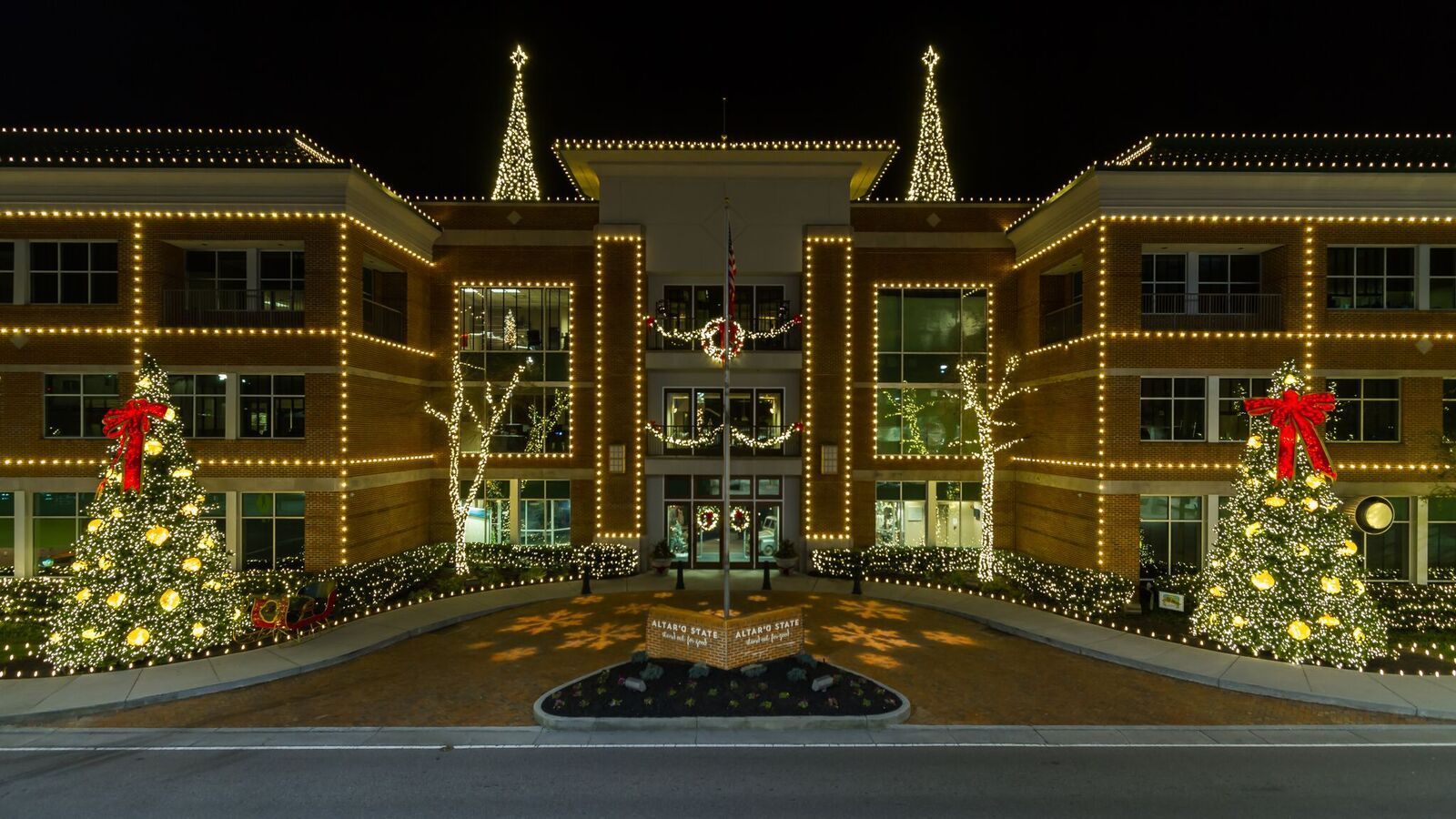 Office building at night, lit with holiday lights; Christmas trees on either side, red bows.