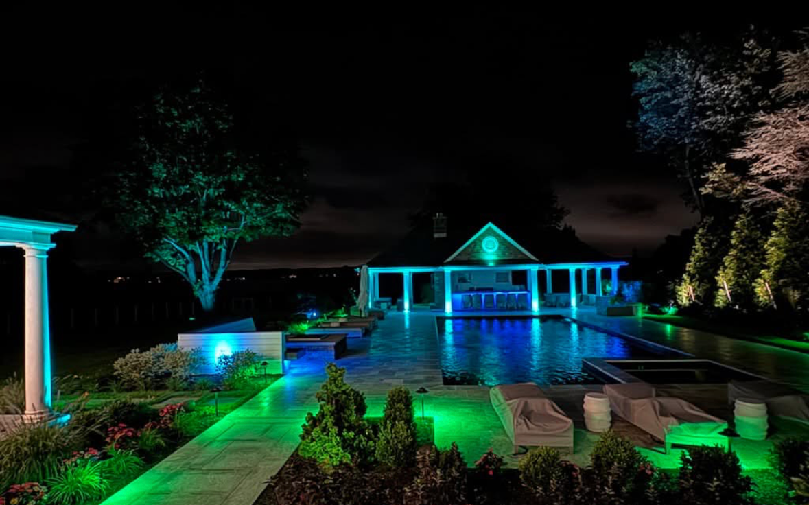 Brick pathway illuminated by spotlights, flanked by shrubs and trees, leading to a building at night.