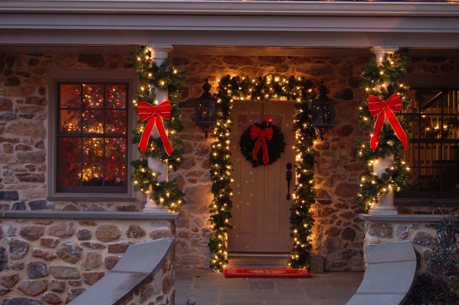 Christmas decorations on a stone house: garlands, lights, red bows, and a wreath on the door.