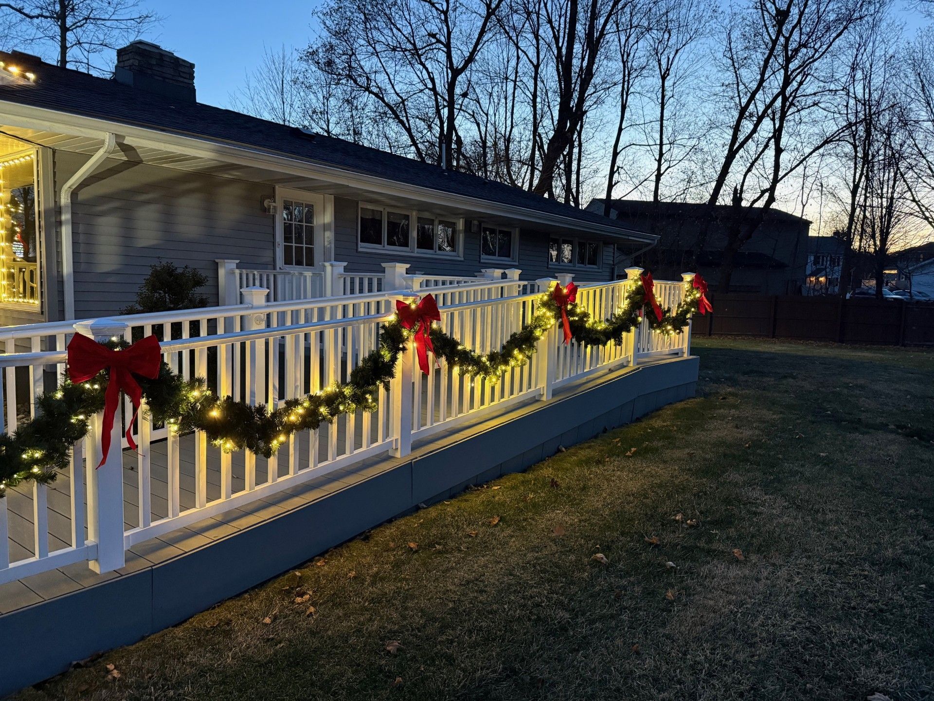 A house decorated with Christmas lights and garland, featuring red bows, on a white railing at dusk.