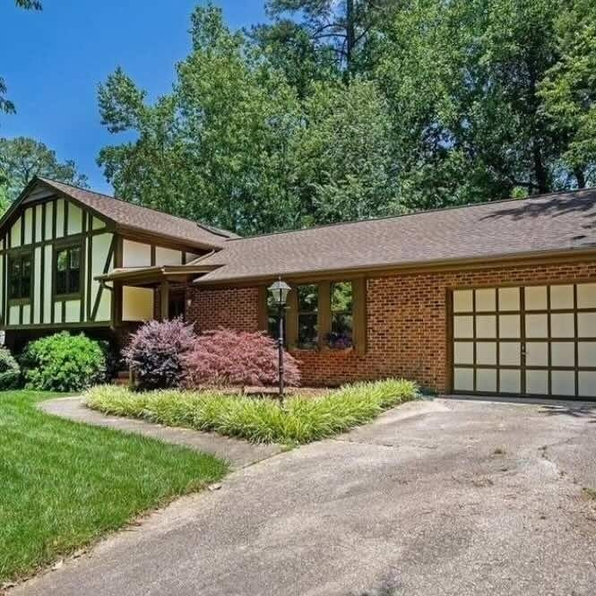 Mid-century brick house with Tudor details, surrounded by greenery and a driveway.