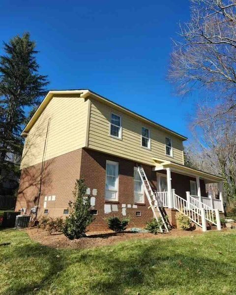 Two-story house with brick base and tan siding. Ladder by porch, blue sky.
