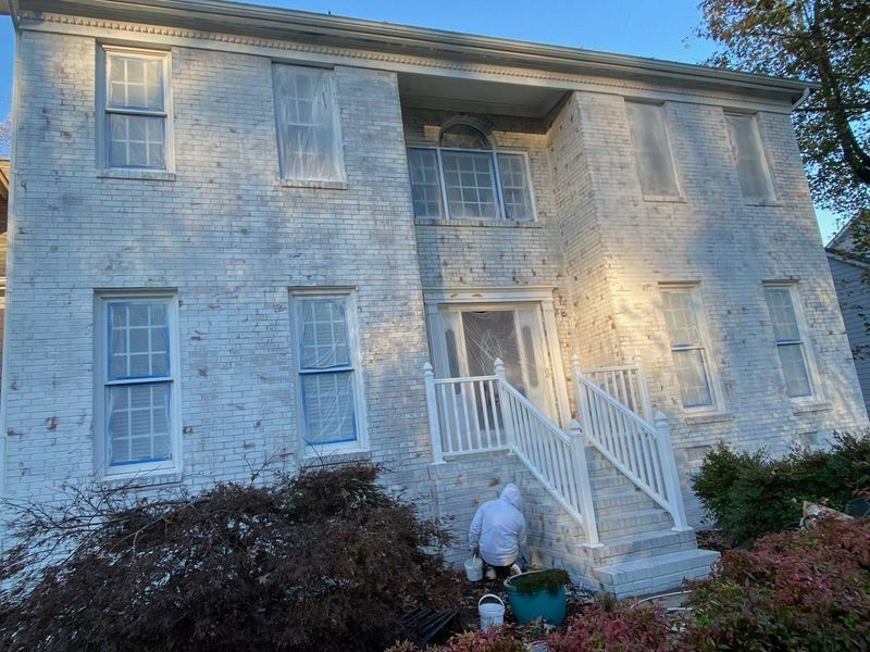 Two-story brick house being painted white. Person in protective suit painting the stairs.