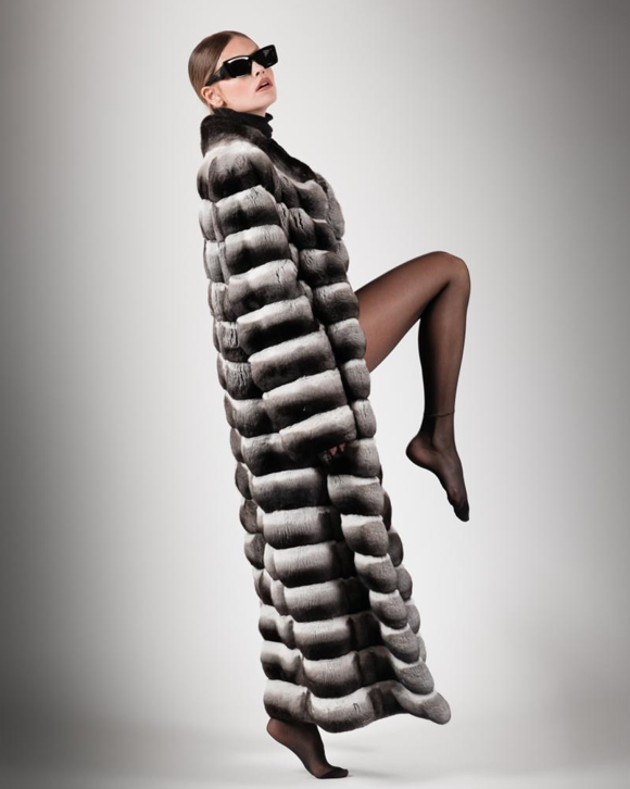 Donna con occhiali da sole e lungo cappotto di pelliccia, una gamba sollevata, su sfondo grigio.