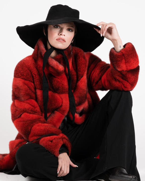 Donna con cappello nero e pelliccia rossa, seduta e che guarda la telecamera.