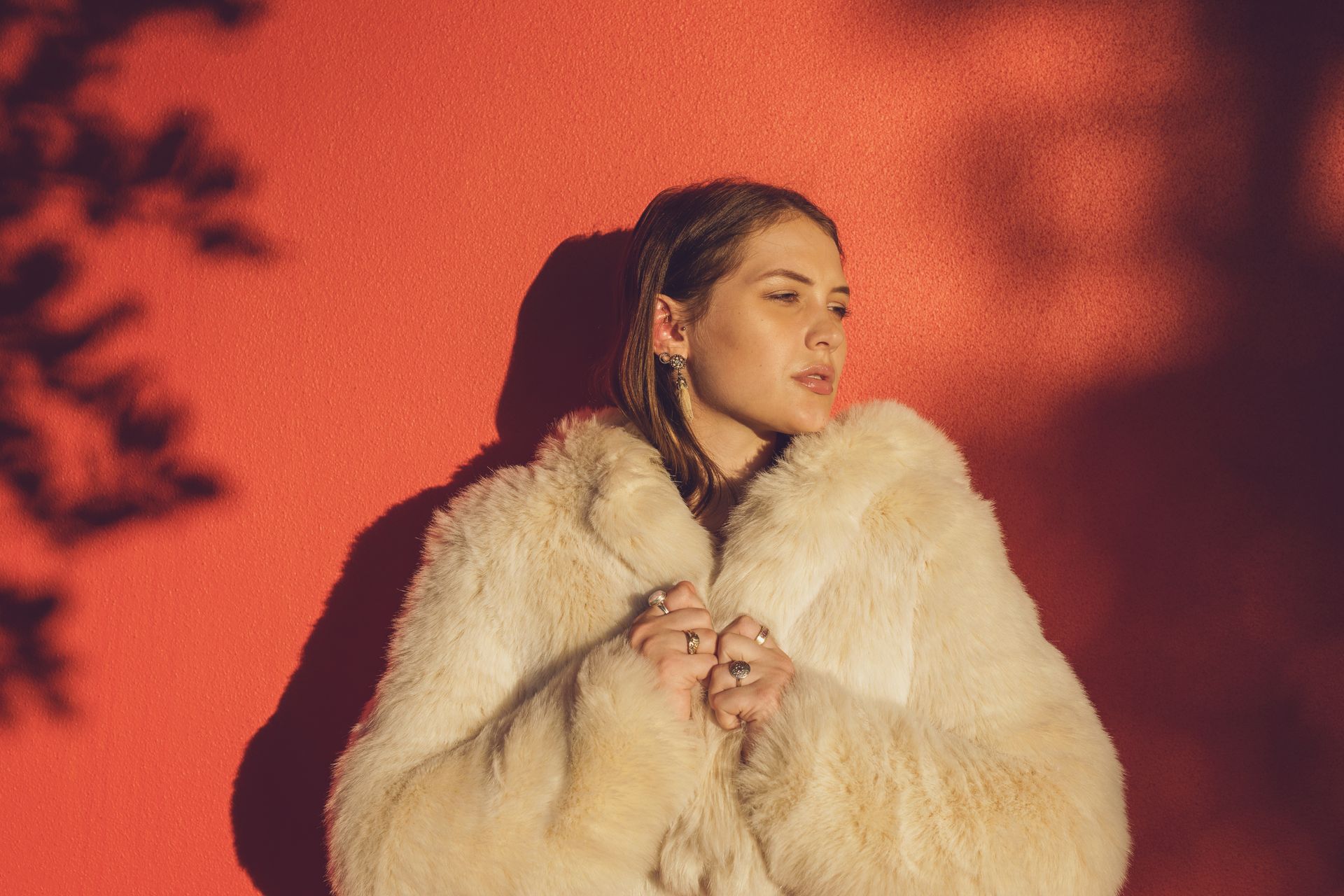 Donna con un soffice camice bianco appoggiata a un muro rosso, guarda di lato. Ombre proiettate nelle vicinanze.