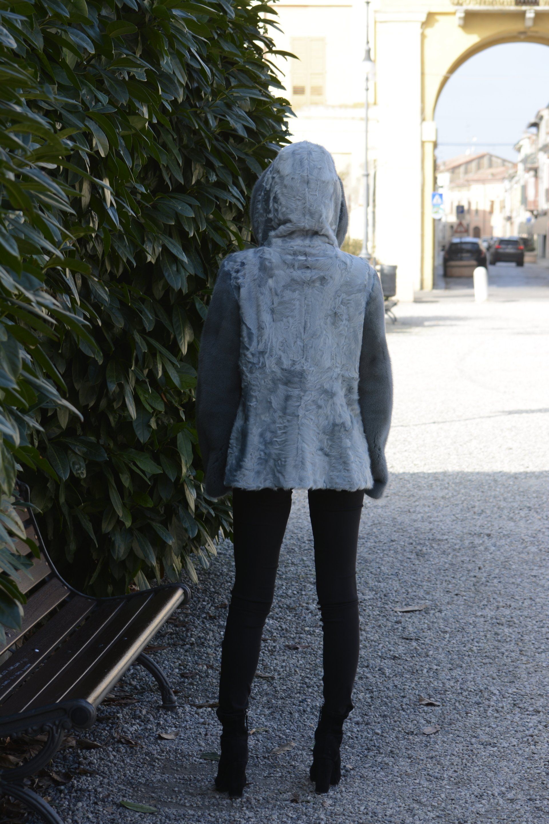 Donna con cappotto grigio e leggings neri in piedi vicino a un cespuglio verde e a una panchina, con una strada e un arco visibili.