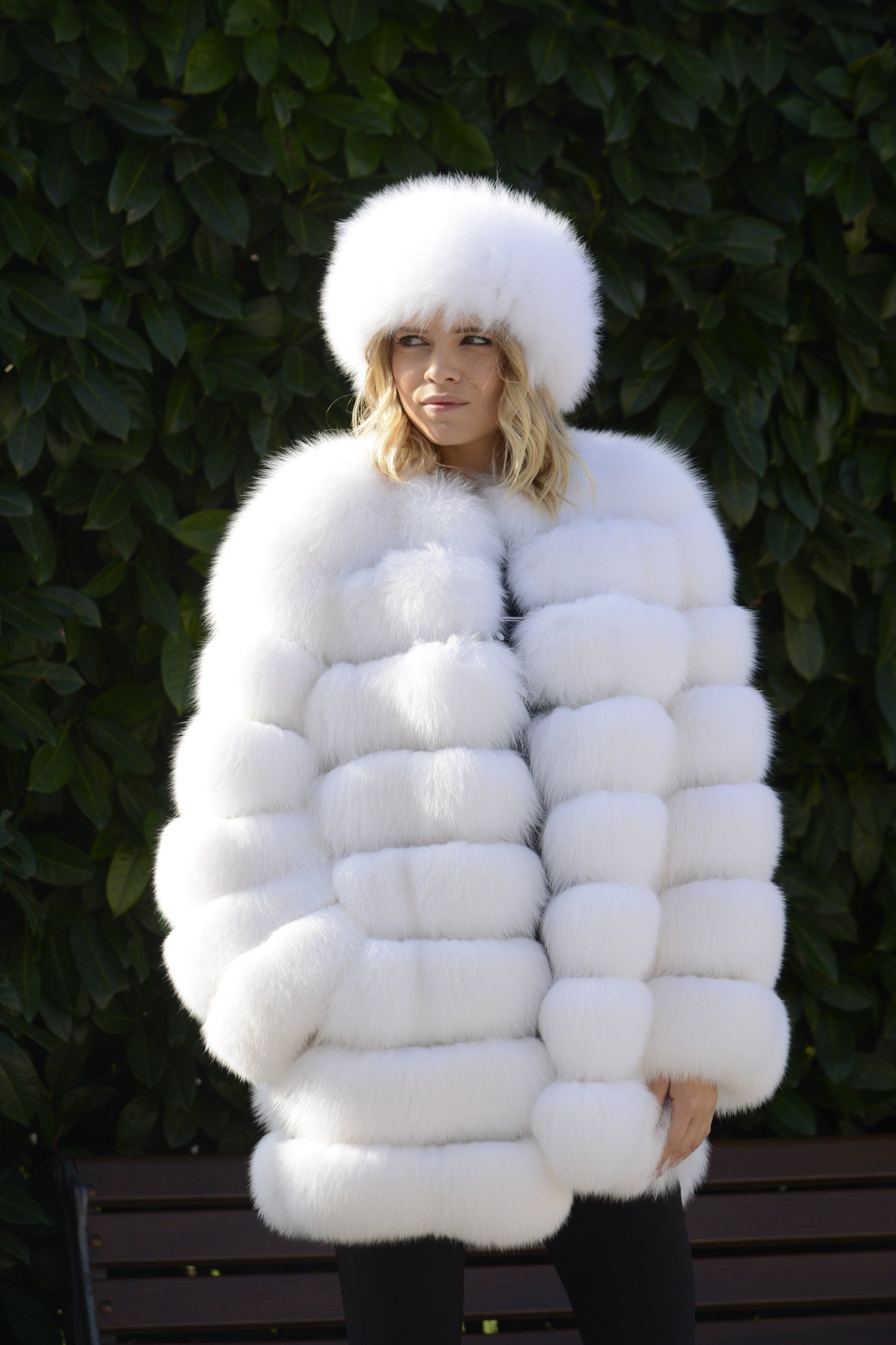 Donna con cappello di pelliccia bianca e attaccapanni all'aperto vicino a una siepe e a una panchina.
