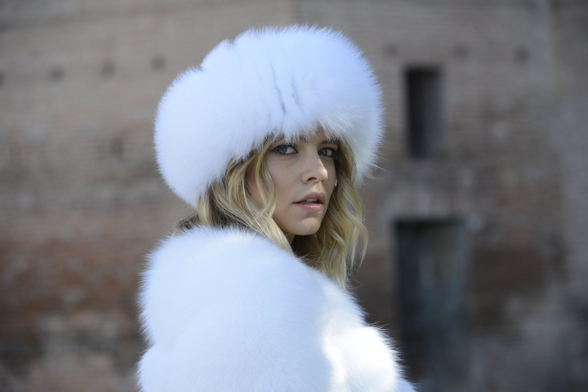 Donna che indossa un cappello di pelliccia bianca e uno scialle, guarda verso la telecamera, su uno sfondo sfocato all'aperto.