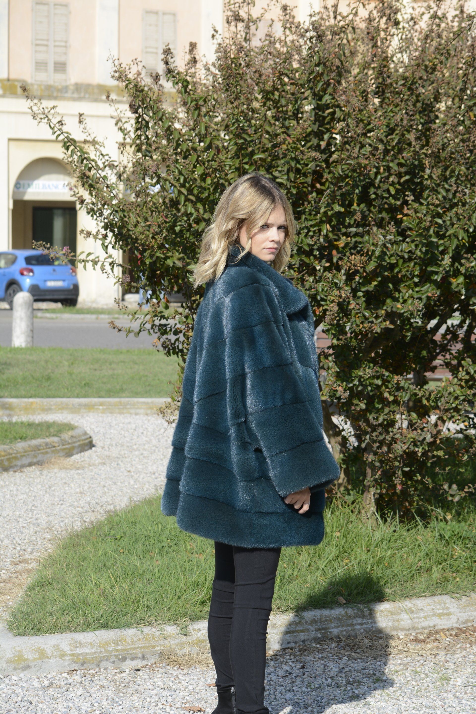 Donna con indosso una pelliccia verde acqua che si guarda alle spalle. In piedi all'aperto, vicino a cespugli verdi e a un edificio.