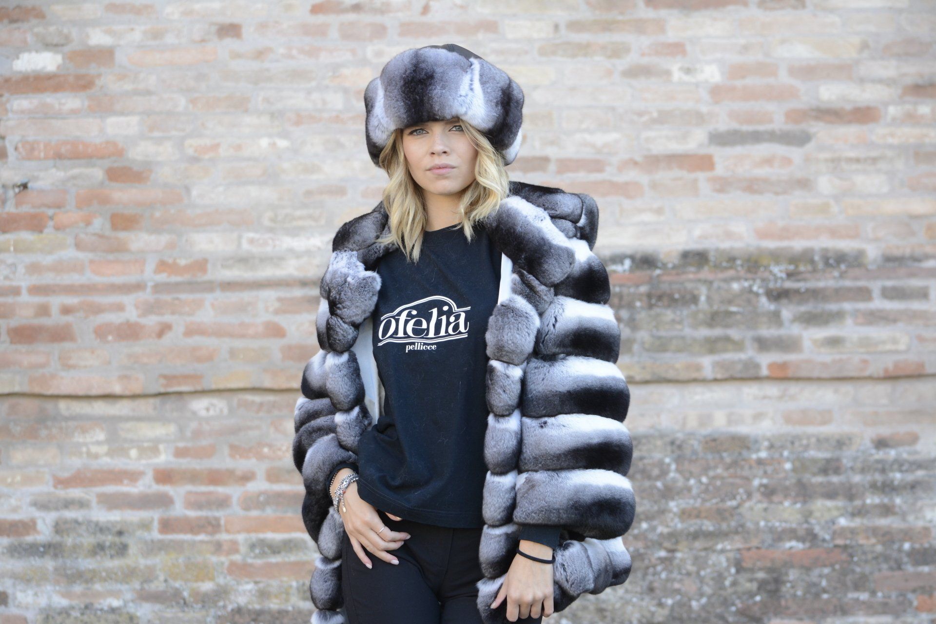Una donna con pelliccia e cappello posa davanti a un muro di mattoni.