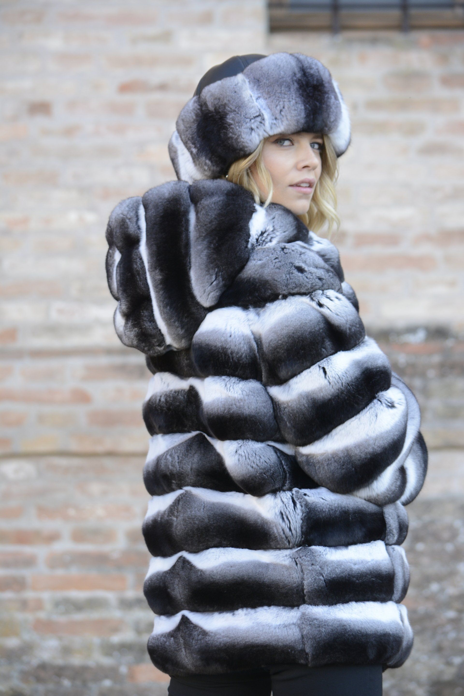 Donna con un cappotto di pelliccia bianco e nero con cappuccio e un cappello, in piedi fuori da un edificio in mattoni.