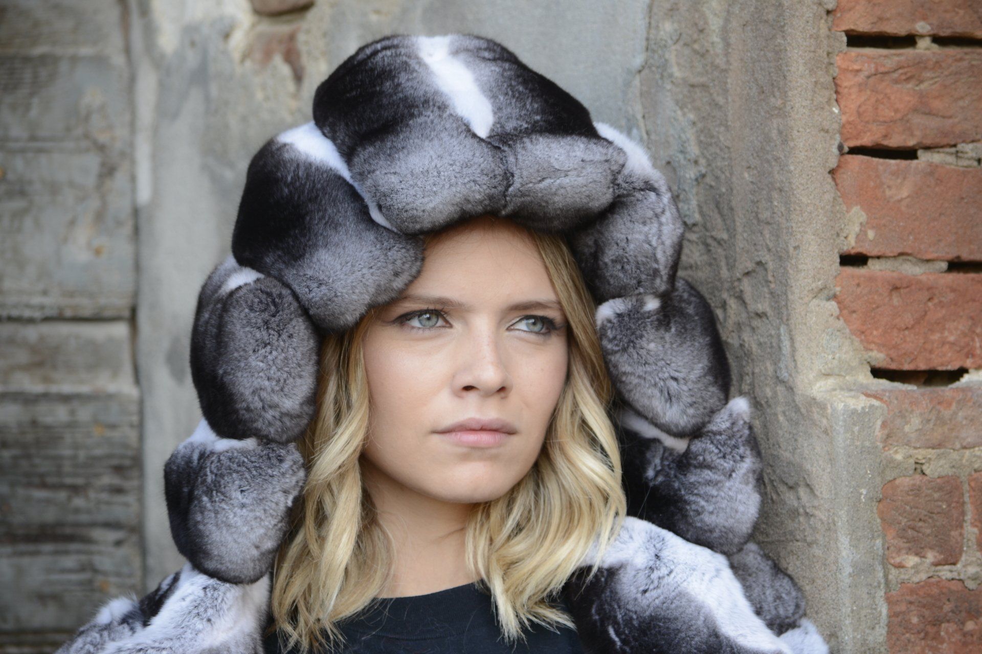 Donna con cappuccio di pelliccia, capelli biondi, in piedi vicino a un muro di mattoni.