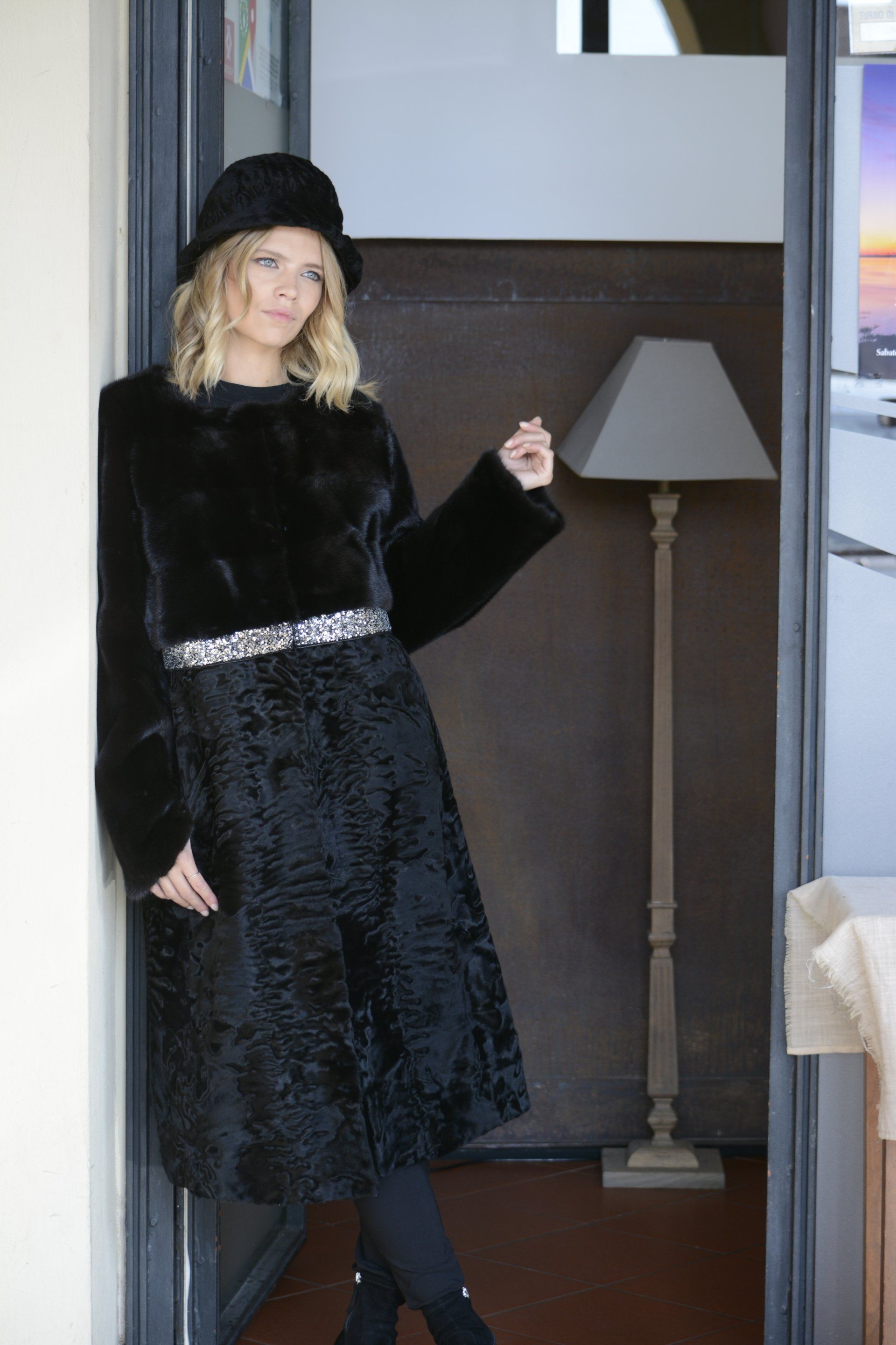 Donna in piedi sulla soglia di una porta, con indosso un cappotto di pelliccia nera, un cappello e una cintura.