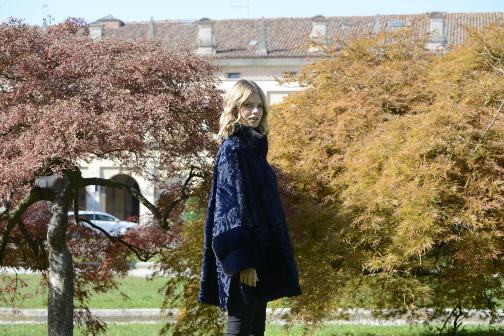 Una donna con un mantello blu scuro è in piedi in un parco con alberi autunnali colorati; sullo sfondo un edificio.