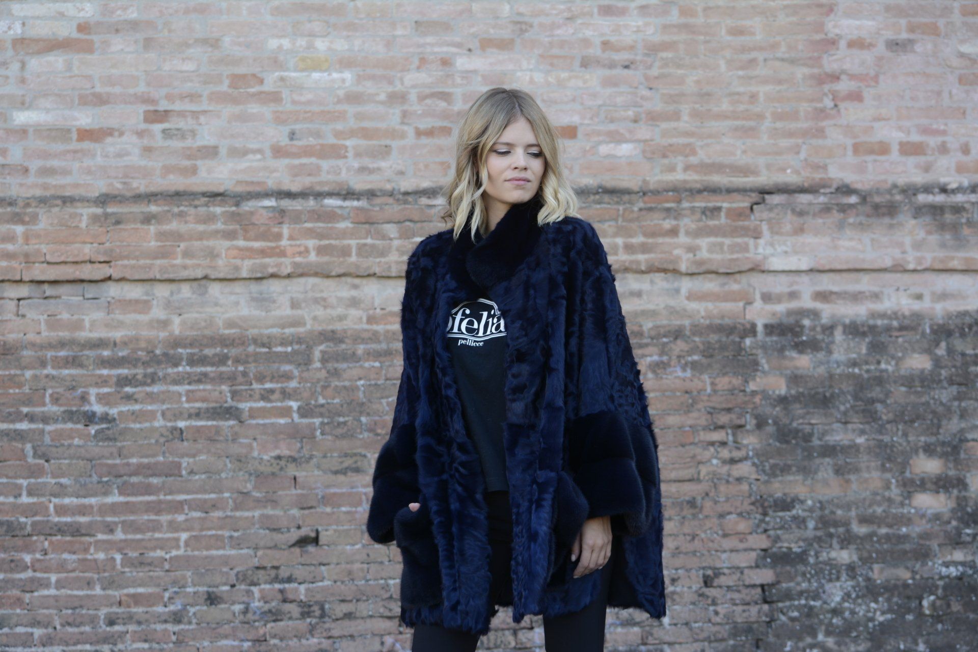 Una donna con cappotto blu scuro, camicia nera e pantaloni è in piedi contro un muro di mattoni.