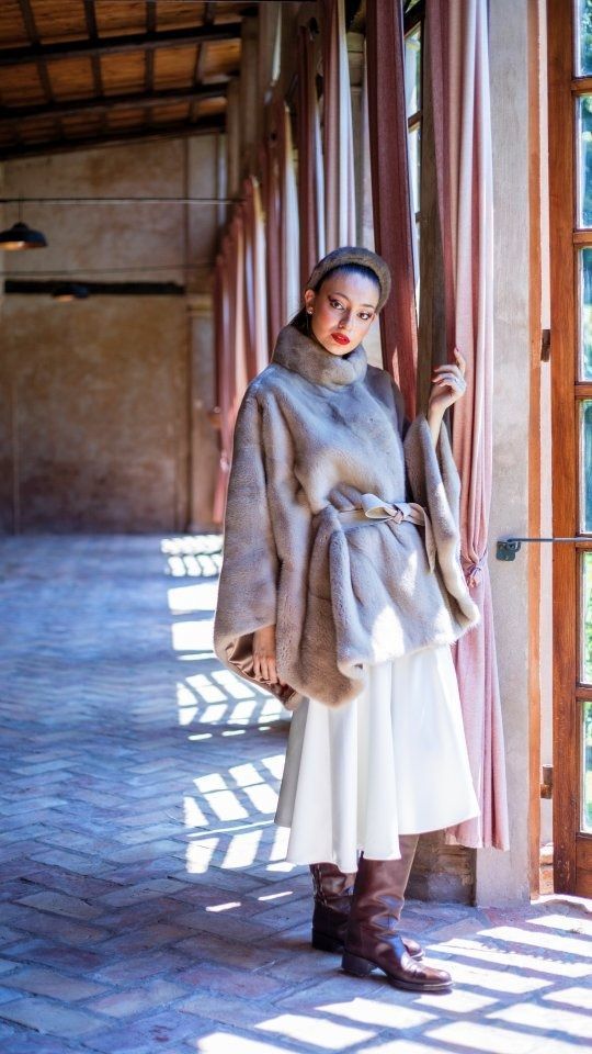 Donna con mantello e gonna beige, stivali marroni, in piedi in un corridoio ad arco con la luce che filtra dalle finestre.