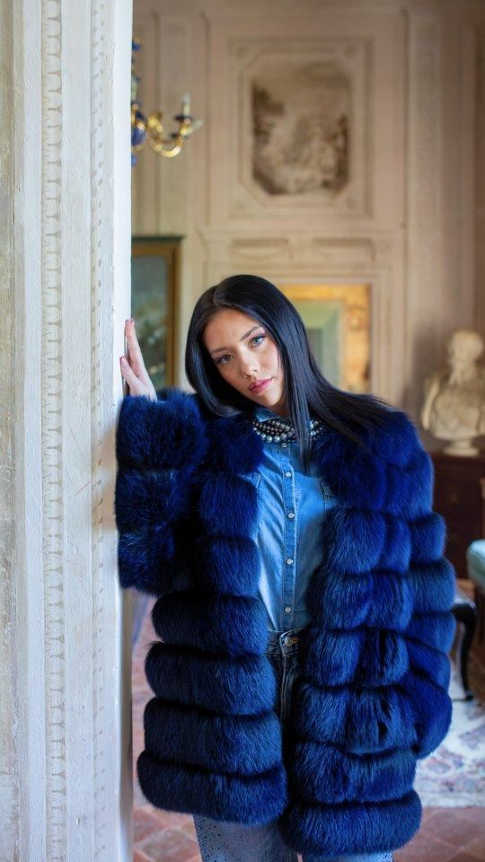 Una donna con una pelliccia blu si appoggia a un pilastro in una stanza decorata.