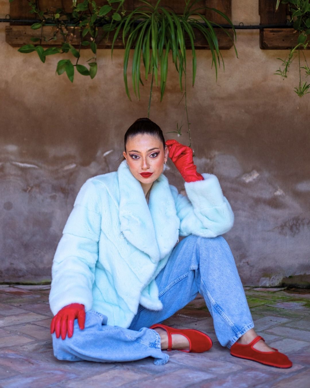 Donna con cappotto azzurro, guanti e scarpe rosse, seduta su un mattone.