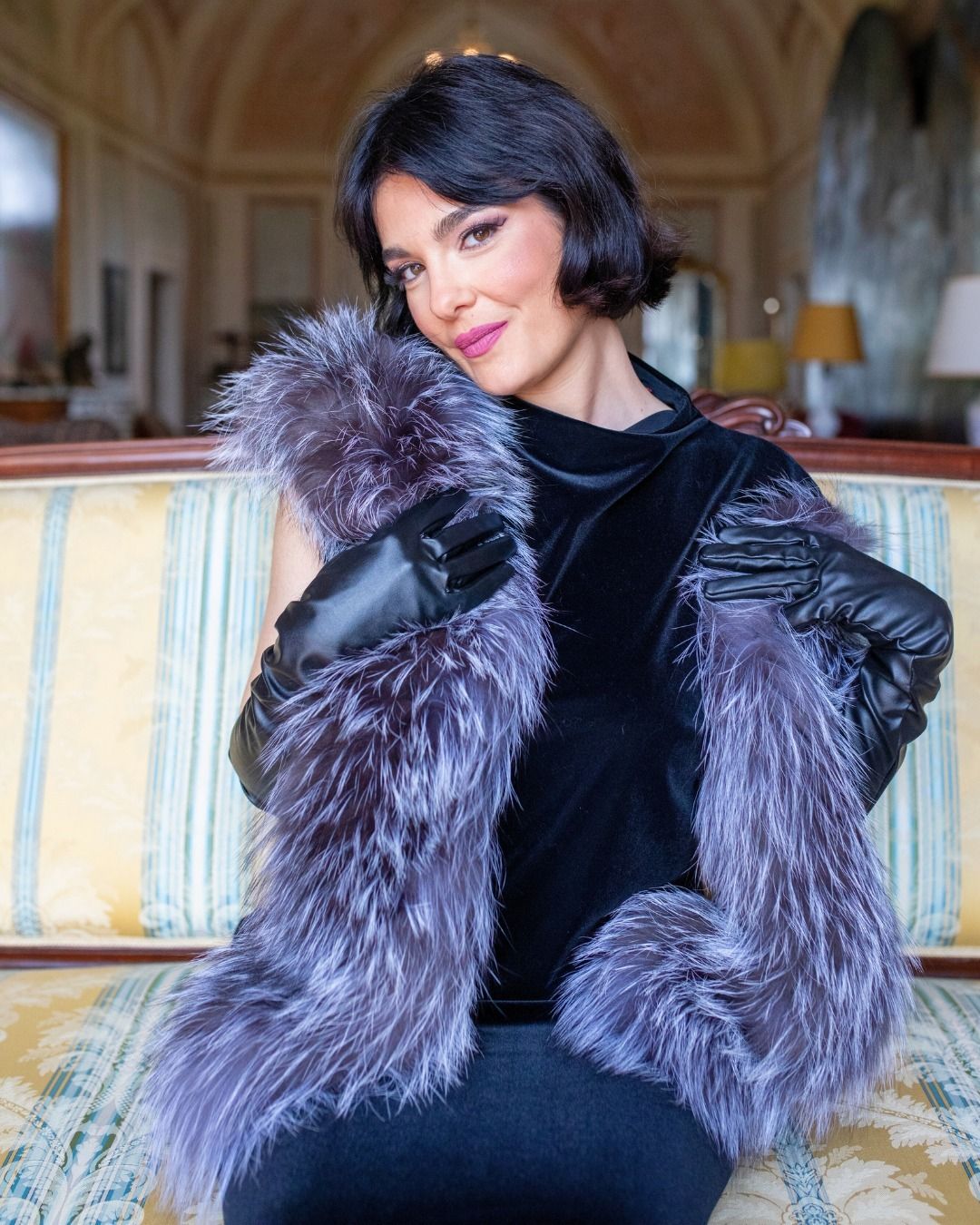 Donna con top di velluto, guanti e stola di pelliccia, seduta su un divano decorato.