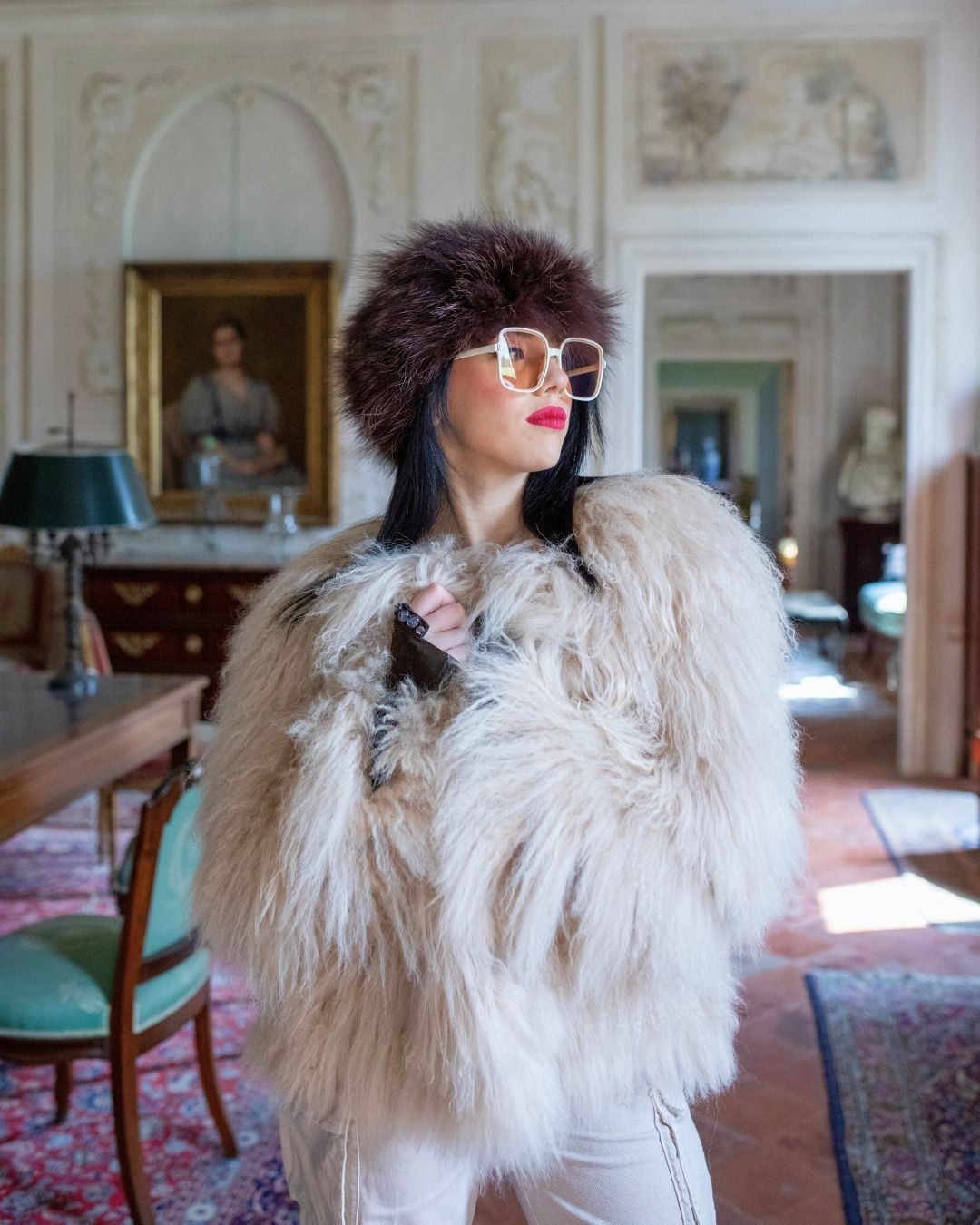 Donna con pelliccia, cappello e occhiali, in una stanza con quadri e mobili antichi.