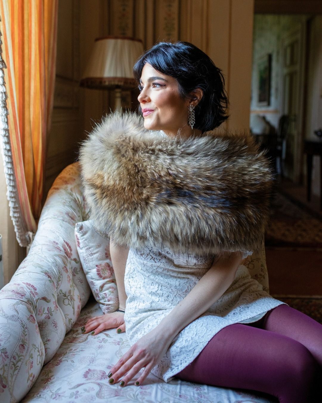 Donna che indossa uno scialle di pelliccia, guarda fuori dalla finestra, seduta su un divano a fiori, con indosso un abito bianco e collant bordeaux.