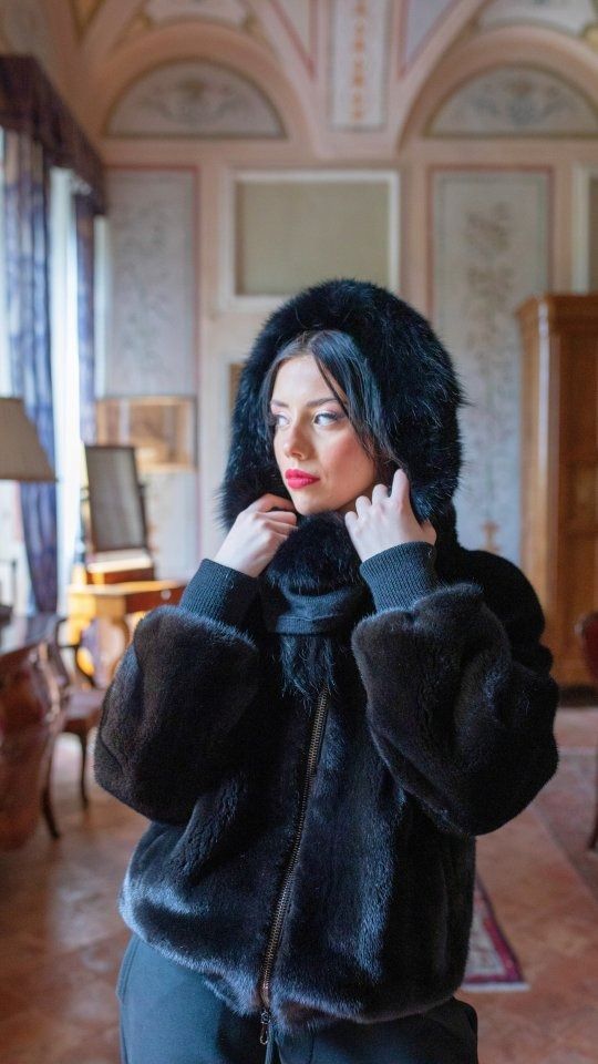 Donna in giacca nera con cappuccio di pelliccia in casa, che guarda altrove con un rossetto rosso.