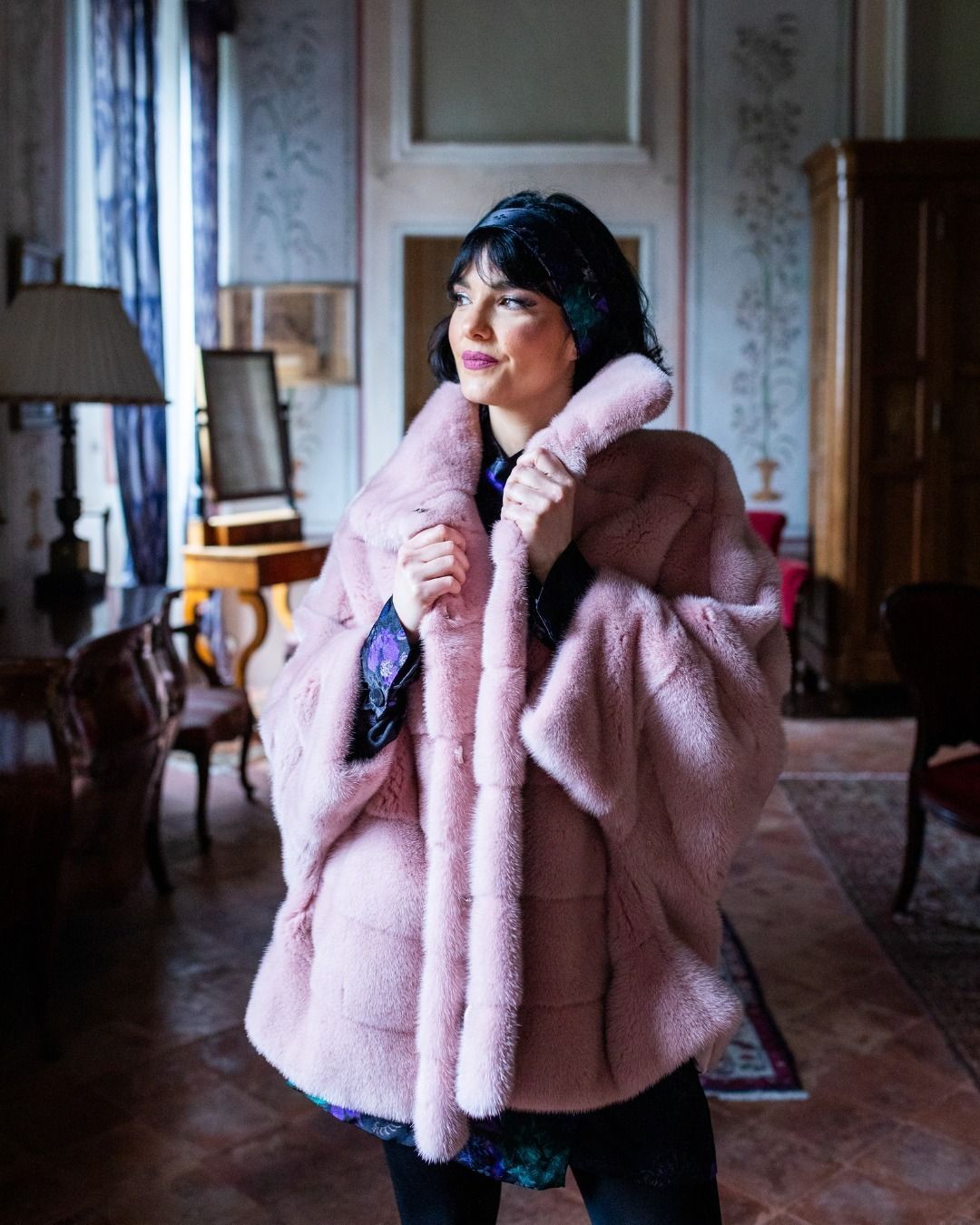 Donna con pelliccia rosa, in piedi in una stanza decorata con mobili in legno scuro e tappeto a motivi geometrici.