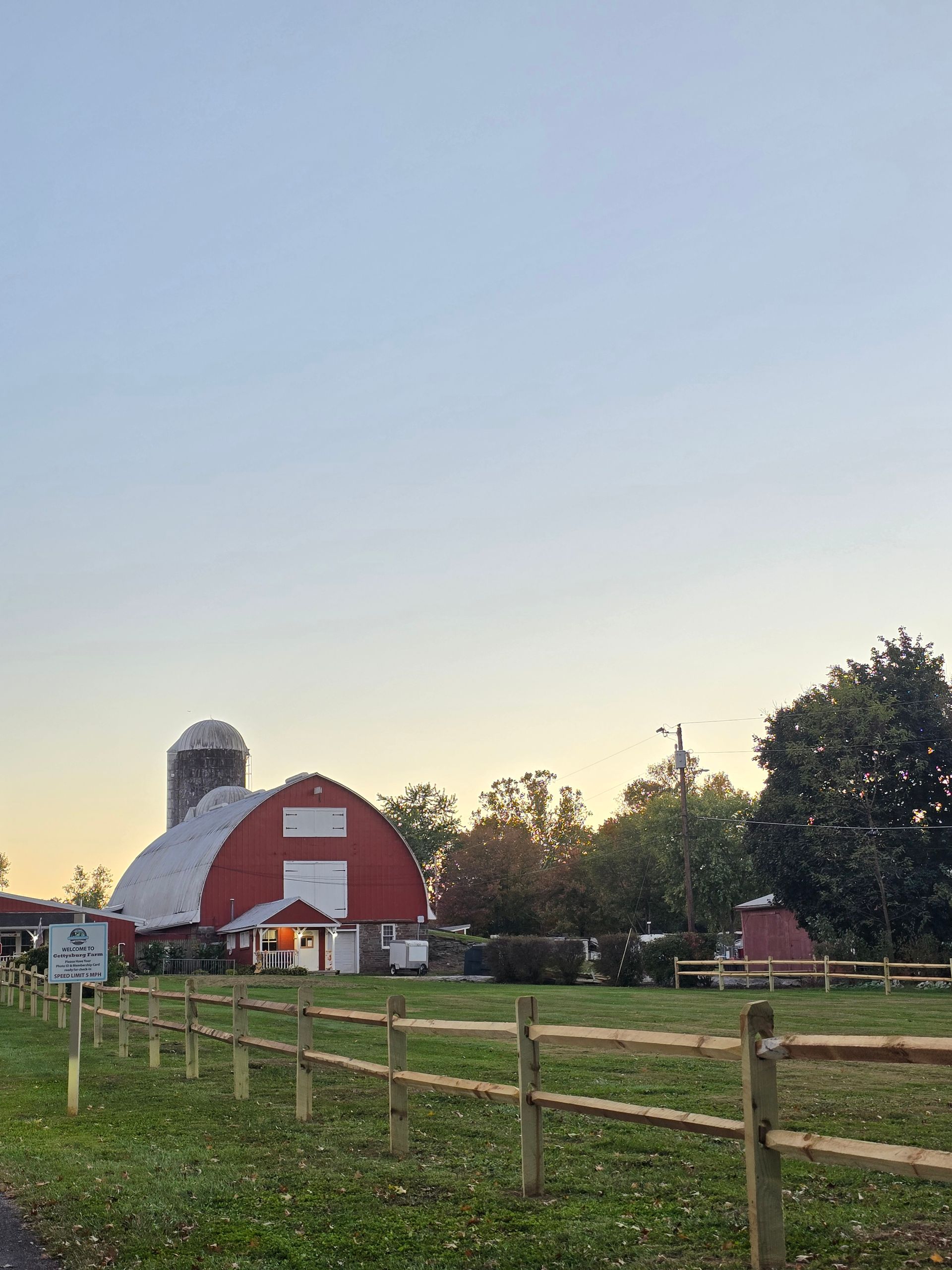 Red barn with silo, wooden fence, and green grass against a light blue sky.