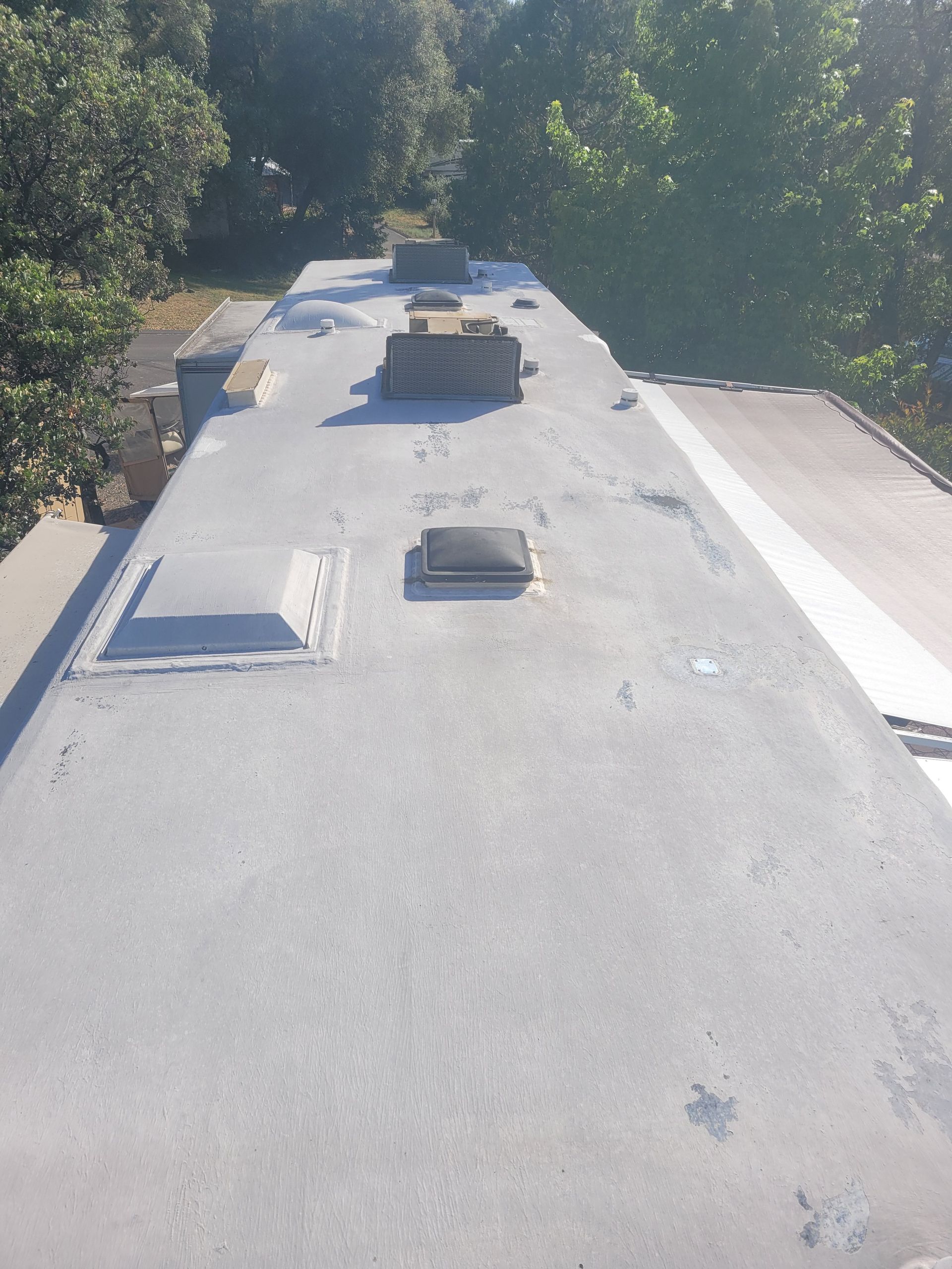 Flat RV rooftop with several vents and HVAC units, bordered by trees in bright sunlight