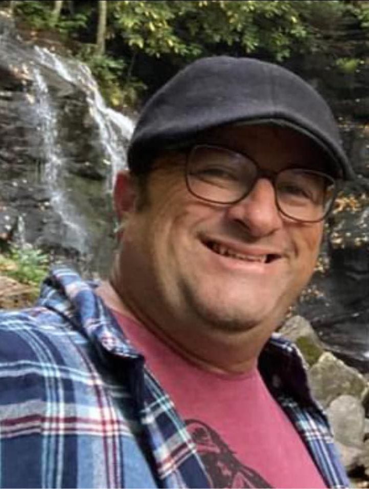A man wearing glasses and a hat is smiling in front of a waterfall.