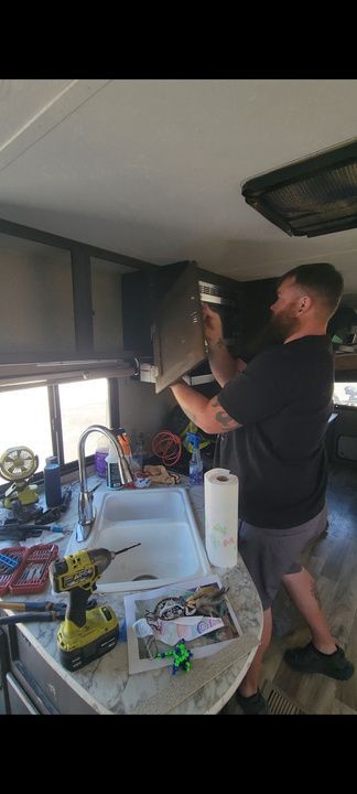 Man working on a cabinet in an RV. He stands by a sink with tools and a paper towel roll.