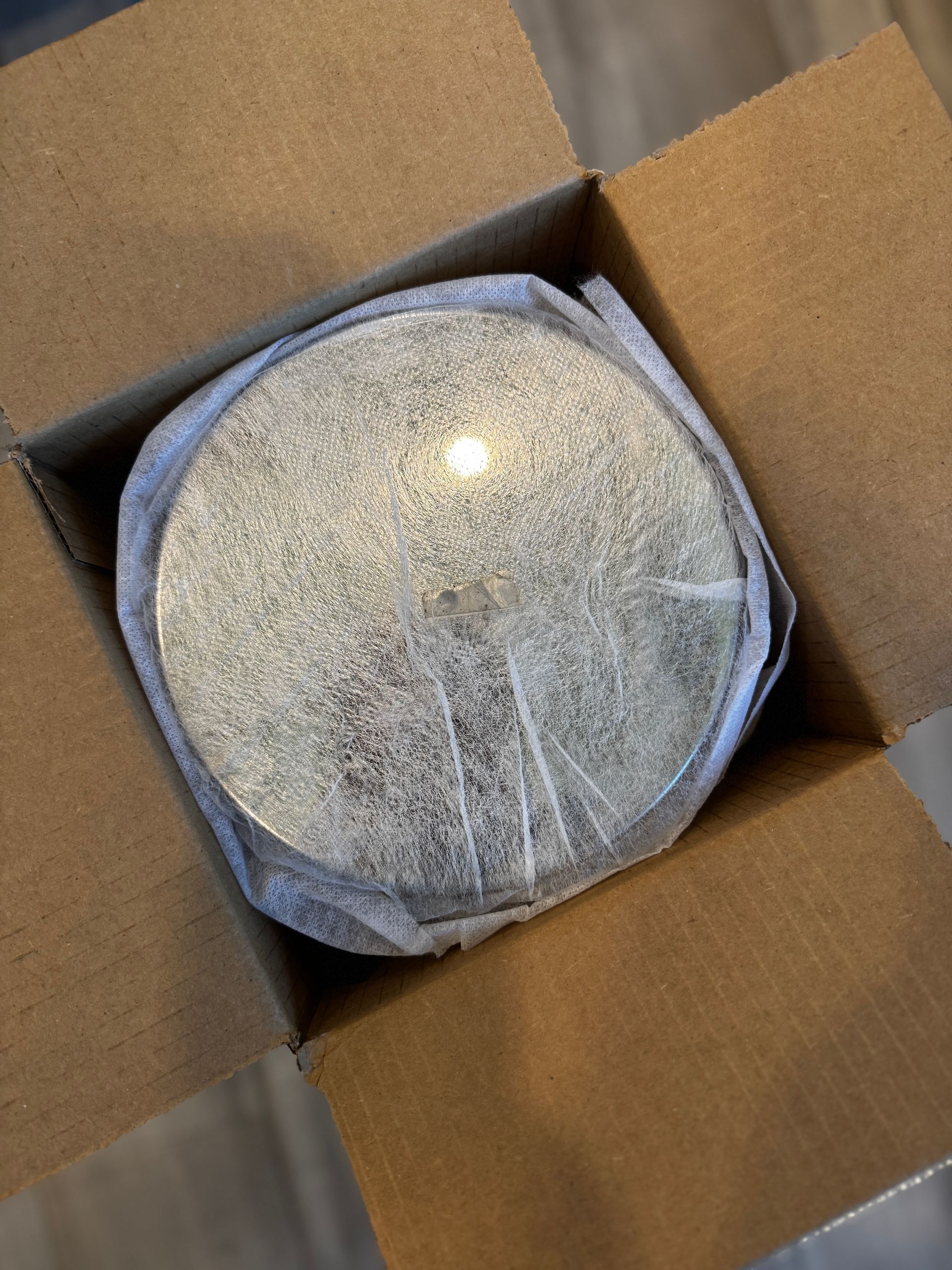 A round, shiny silver object wrapped in white packaging inside an open cardboard box.