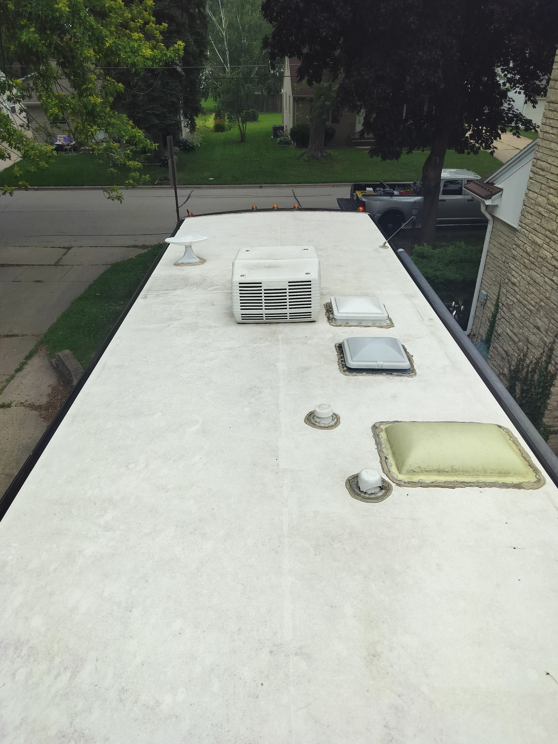An elevated view of a white, weathered RV roof with an air conditioner, skylights, and vents, seen from above a driveway.