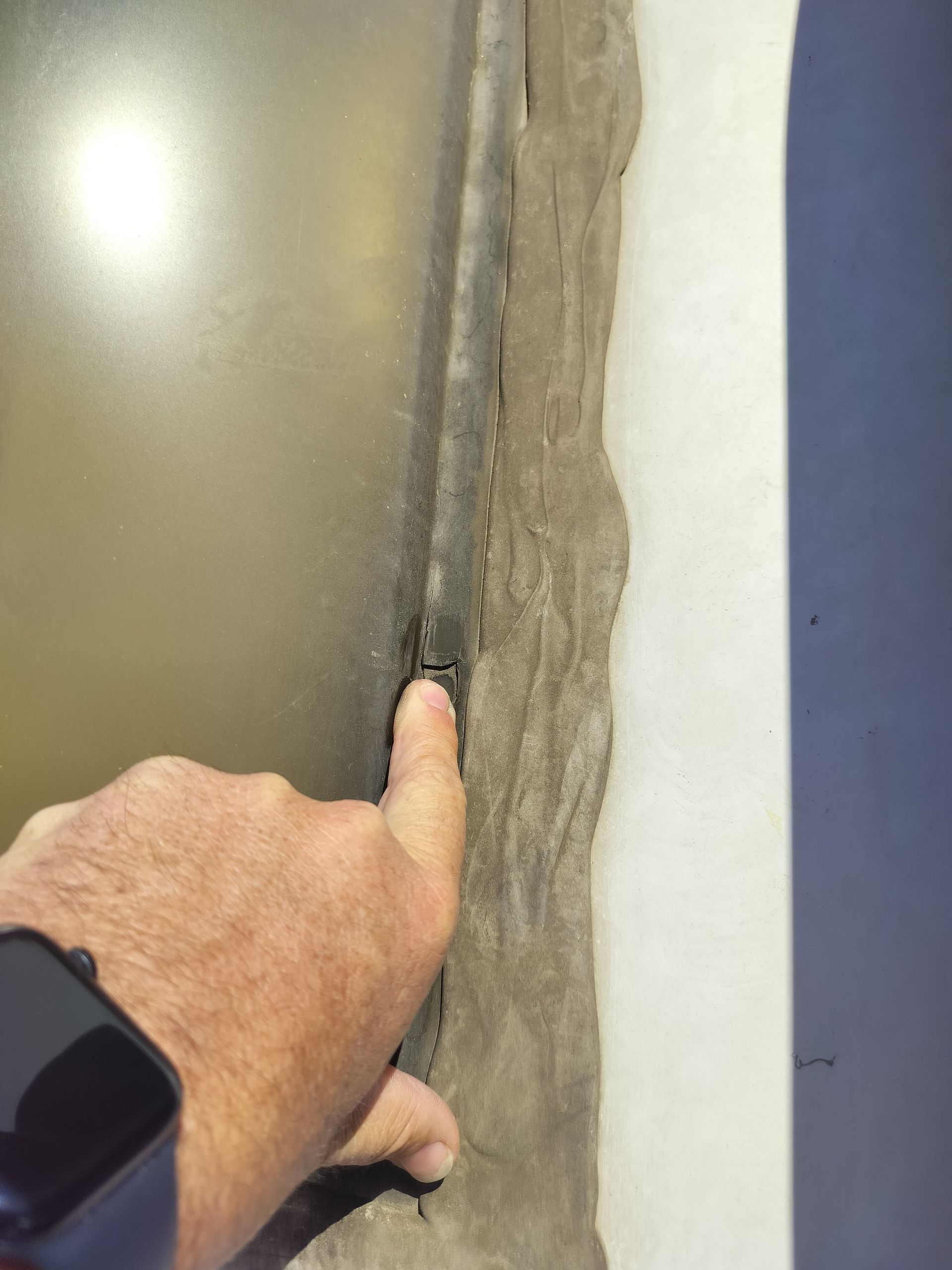 A person's finger points to a gap in the weathered, uneven sealant around an exterior skylight frame on the RV roof.