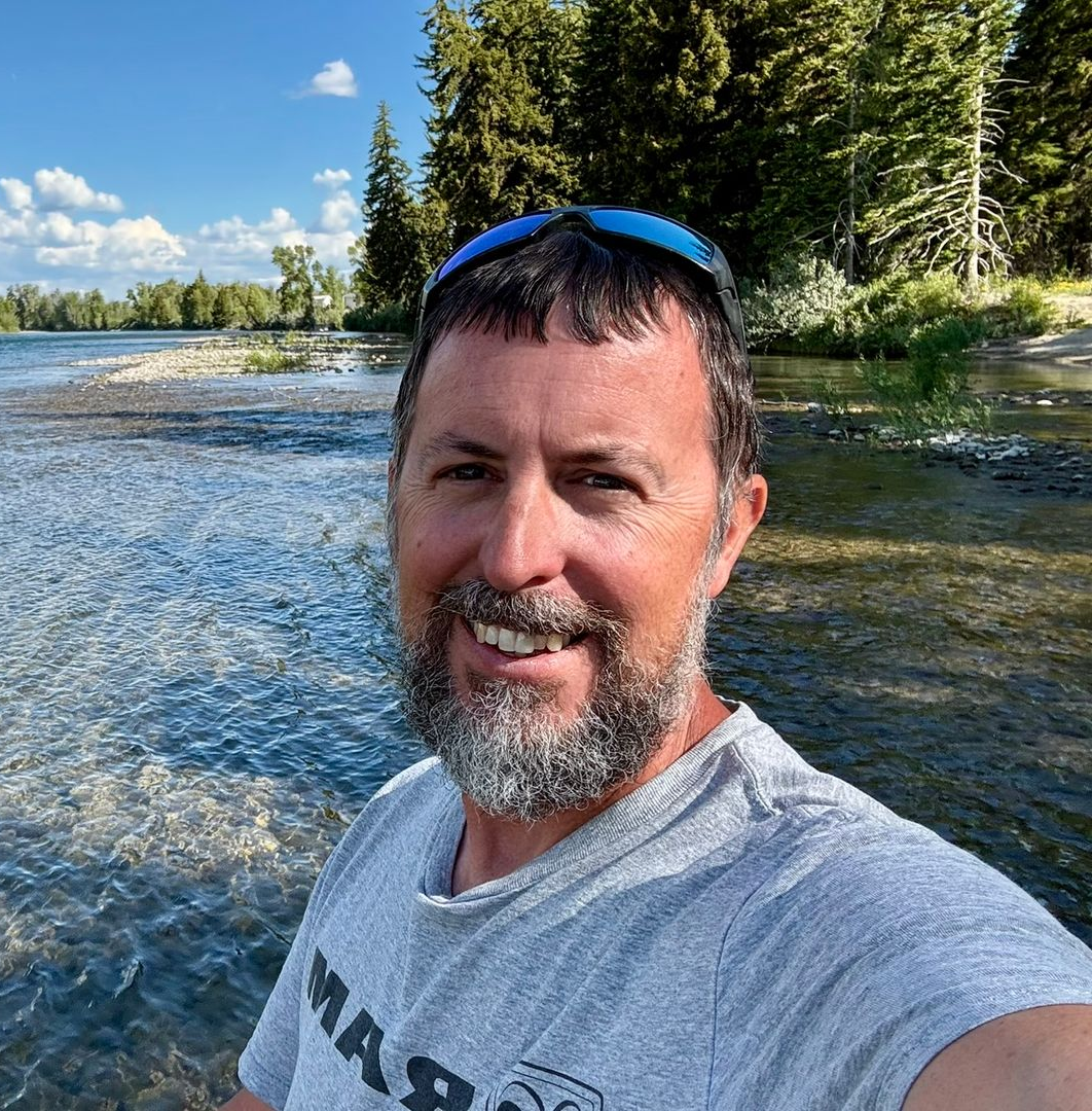 Man with beard standing in front of Lake with sunglasses on his head.