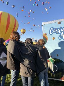 A group of people are standing in front of a trailer with hot air balloons flying in the sky.
