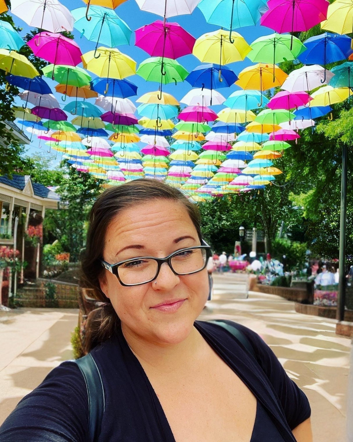A woman wearing glasses takes a selfie under a bunch of colorful umbrellas