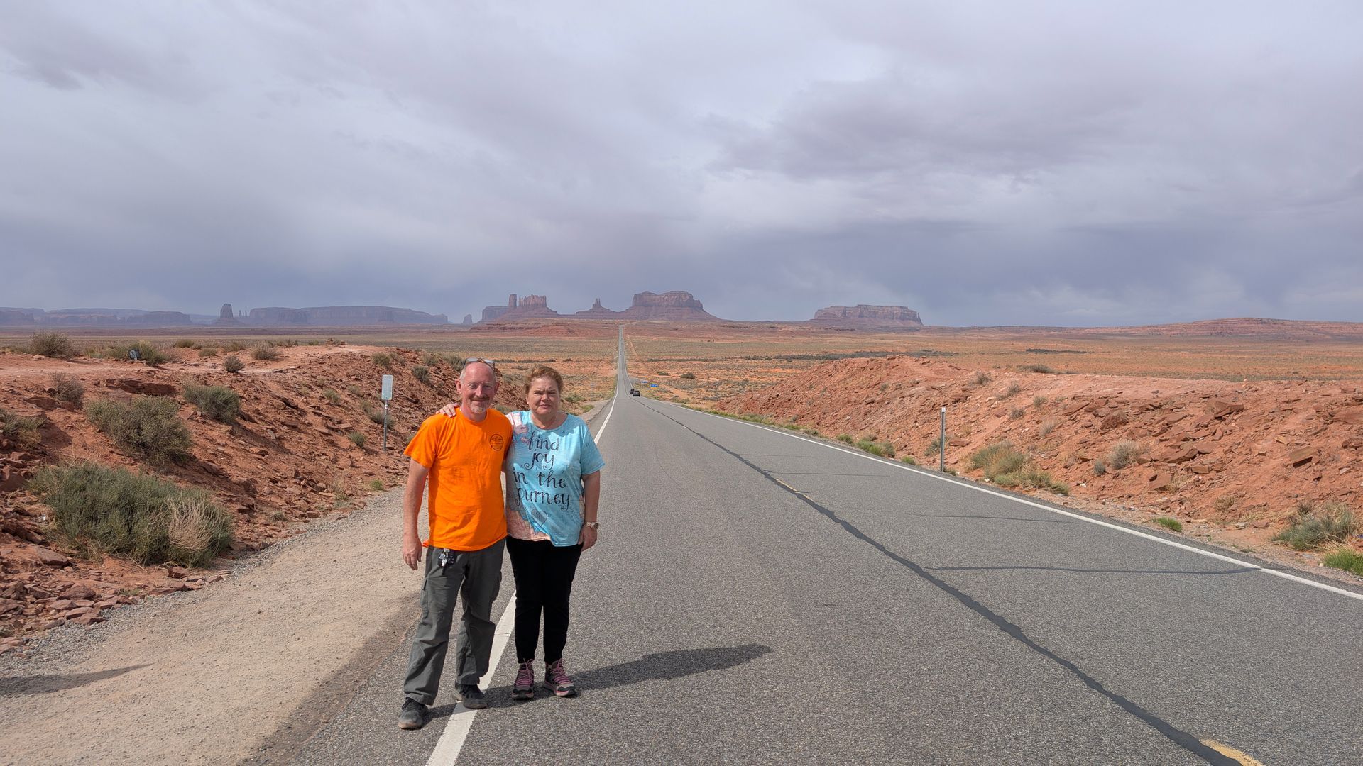 Two people stand on a desert road with red rock formations in the distance under a cloudy sky.