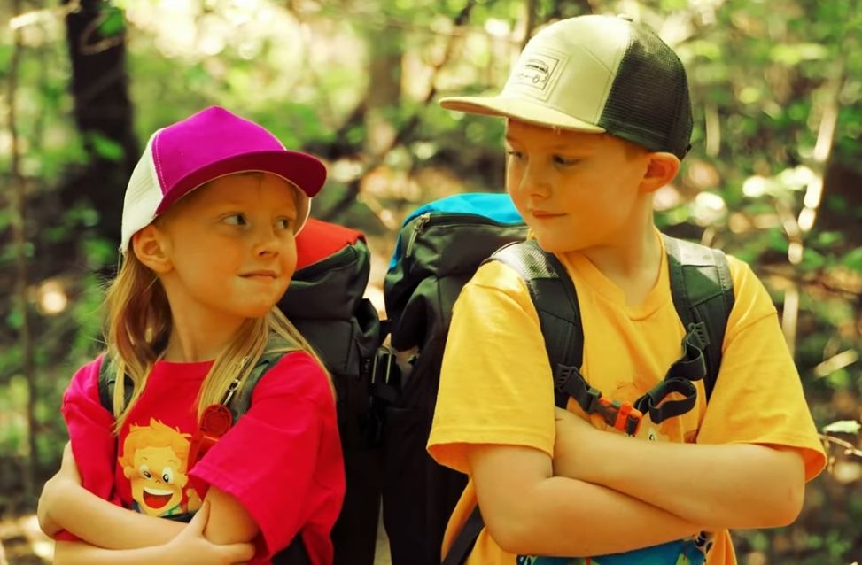 Two children in caps and backpacks standing in a forest, looking at each other with arms crossed.